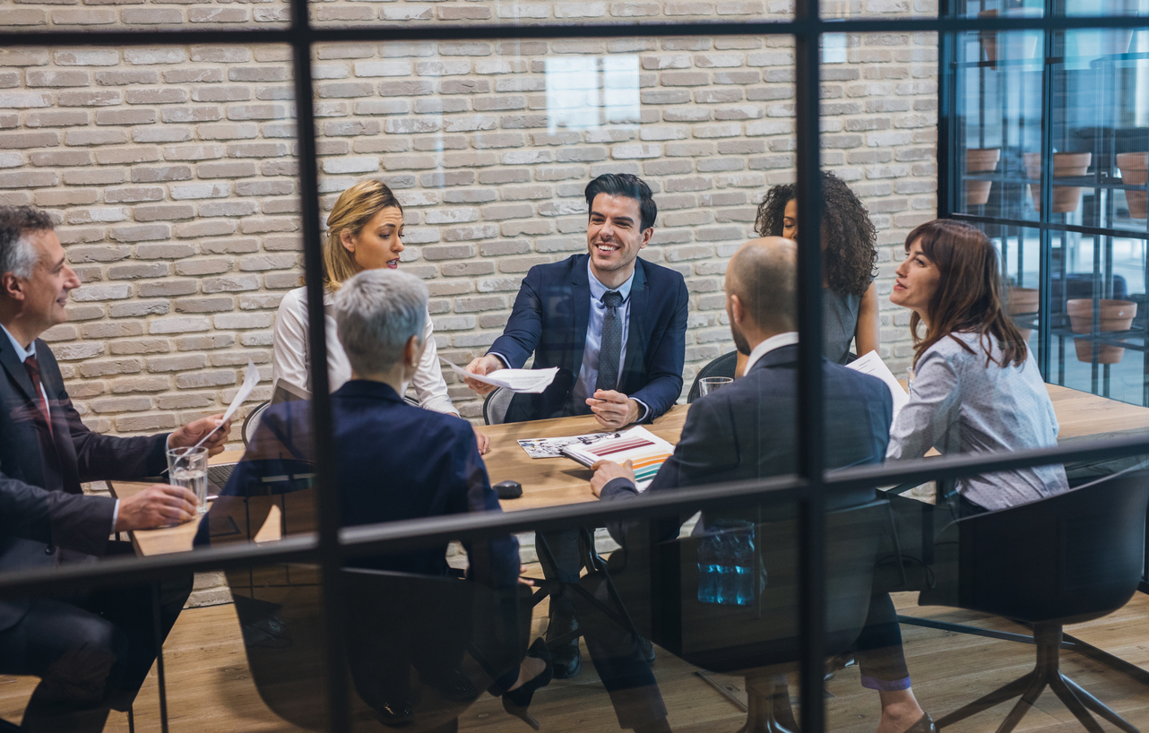 A deal team in discussion around a conference table, seen through the glass of the meeting room